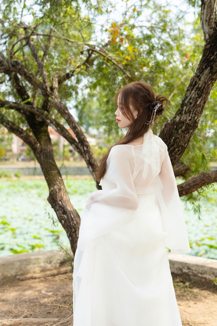 Bride in flowing white dress, back view, near a serene lakeside setting with trees.