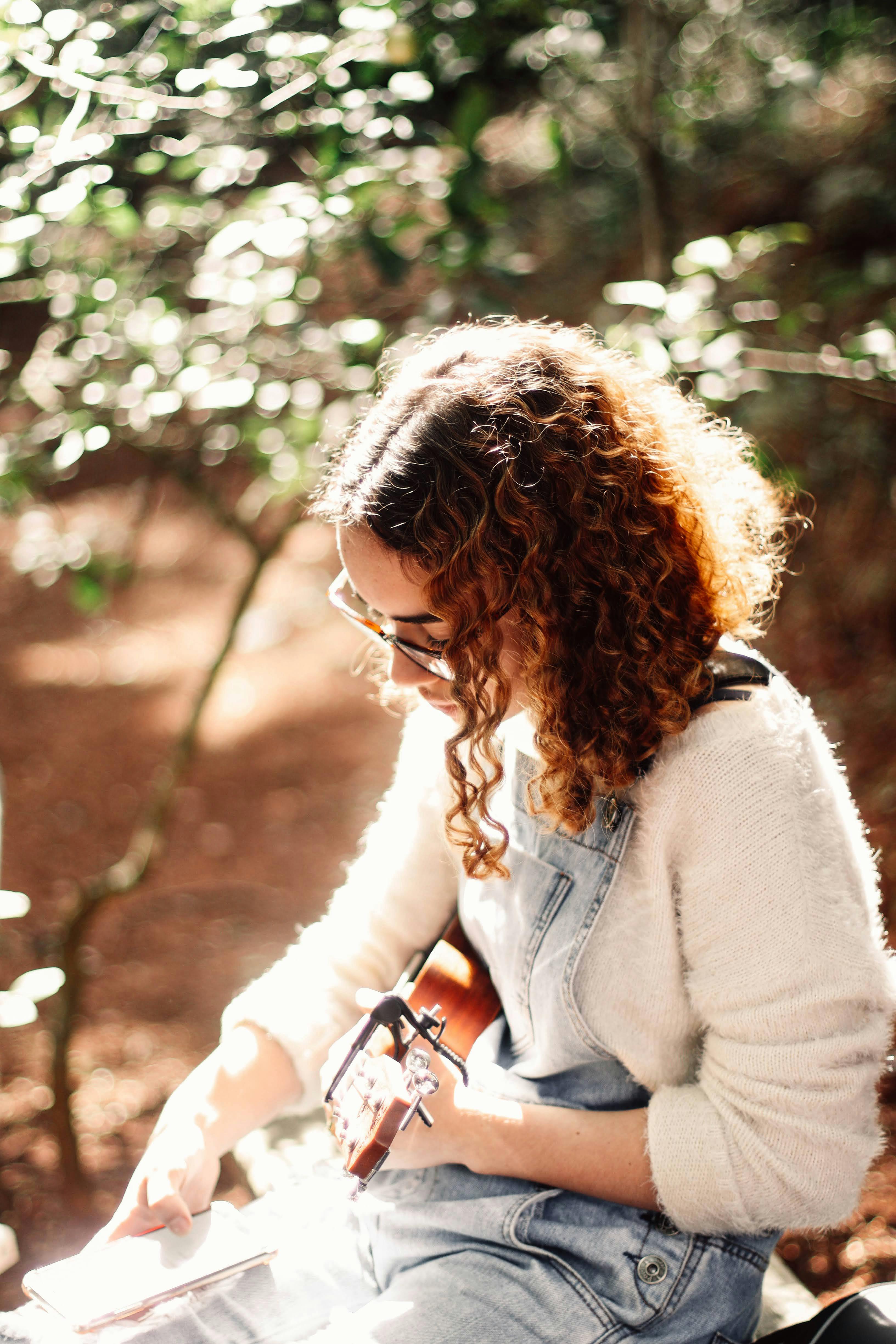 Free A young woman with curly hair playing the ukulele outside in a sun-dappled forest setting. Stock Photo
