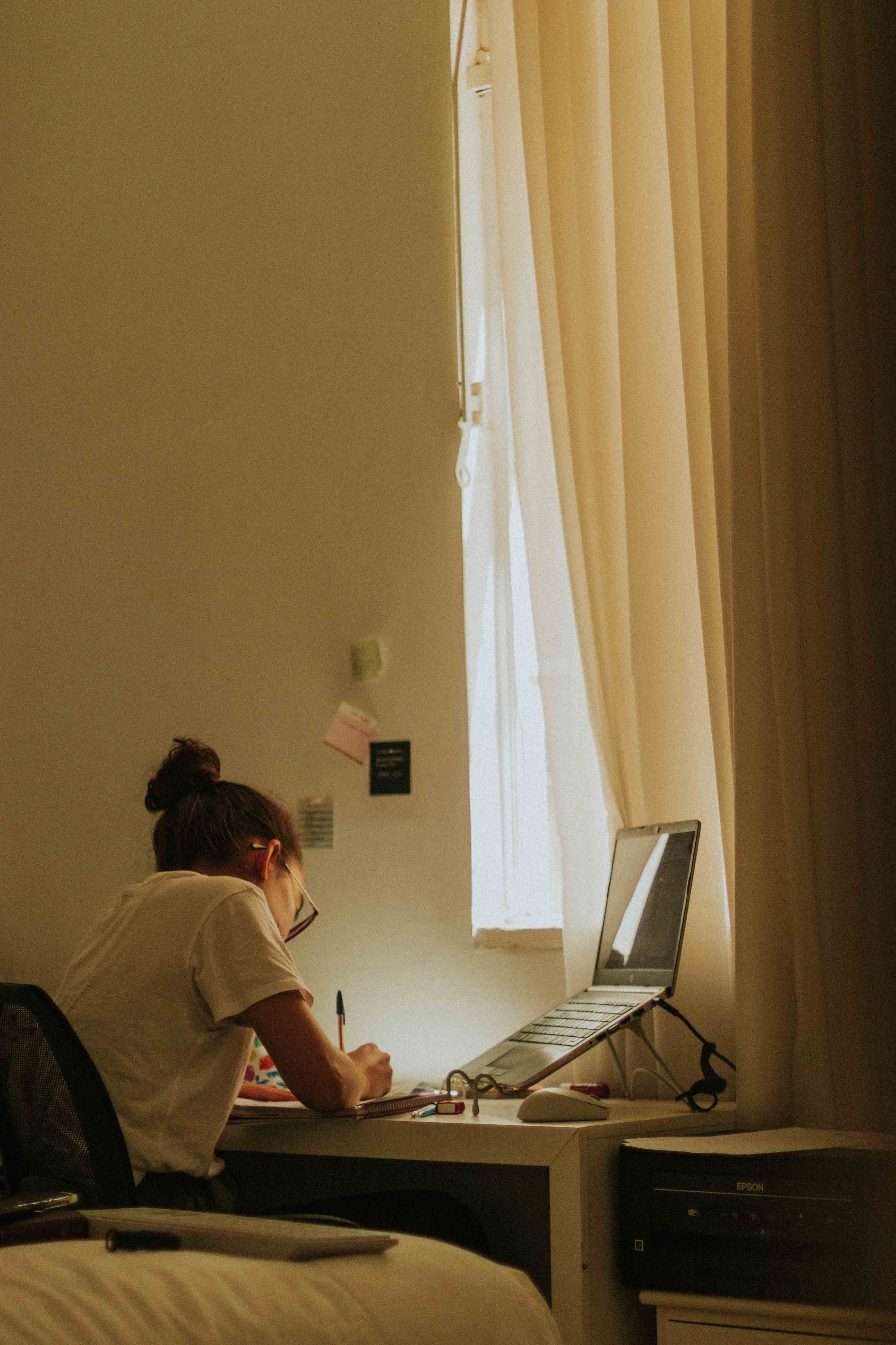A desk with a notebook and laptop near a window