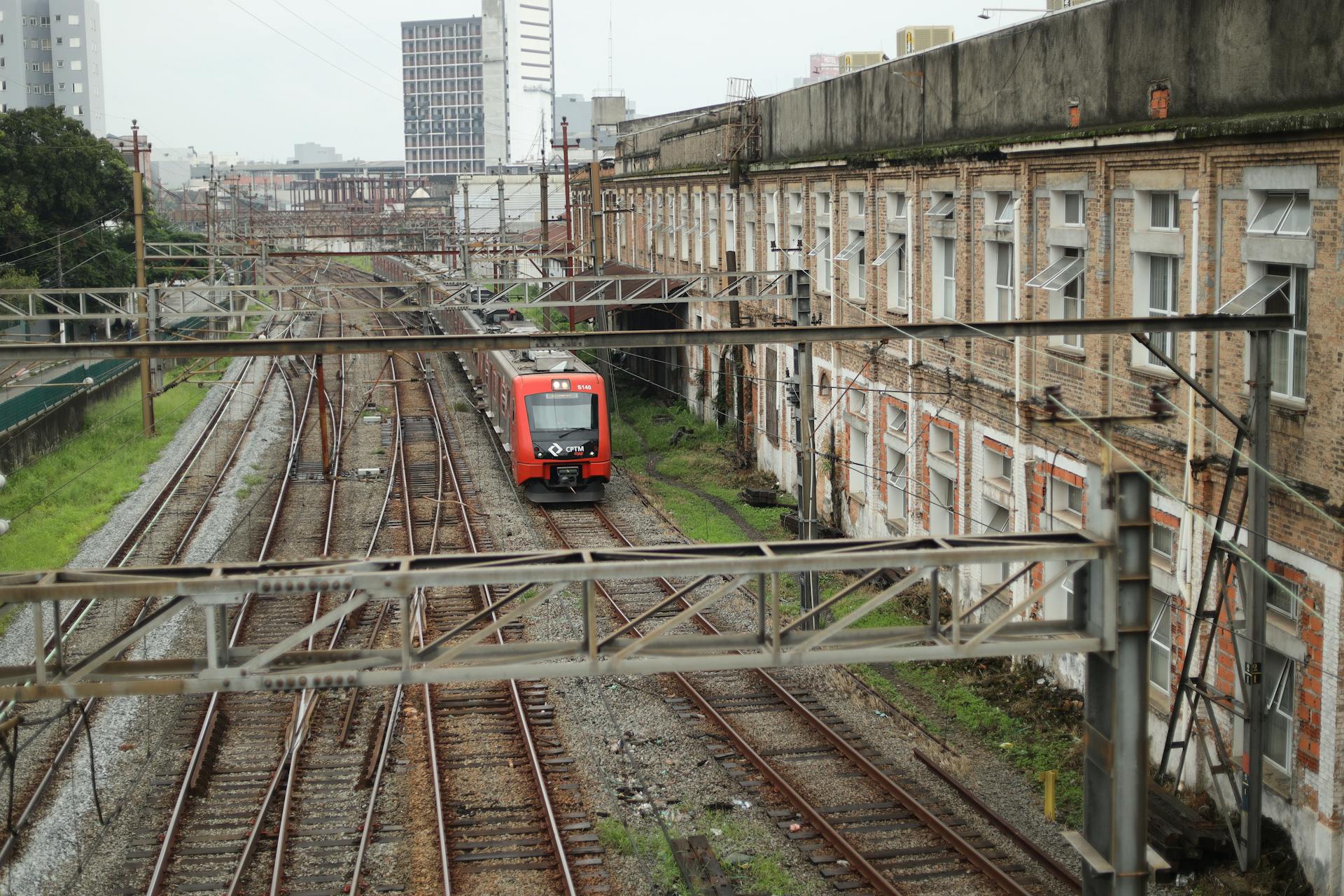 Red train travels along urban railway track beside industrial buildings.