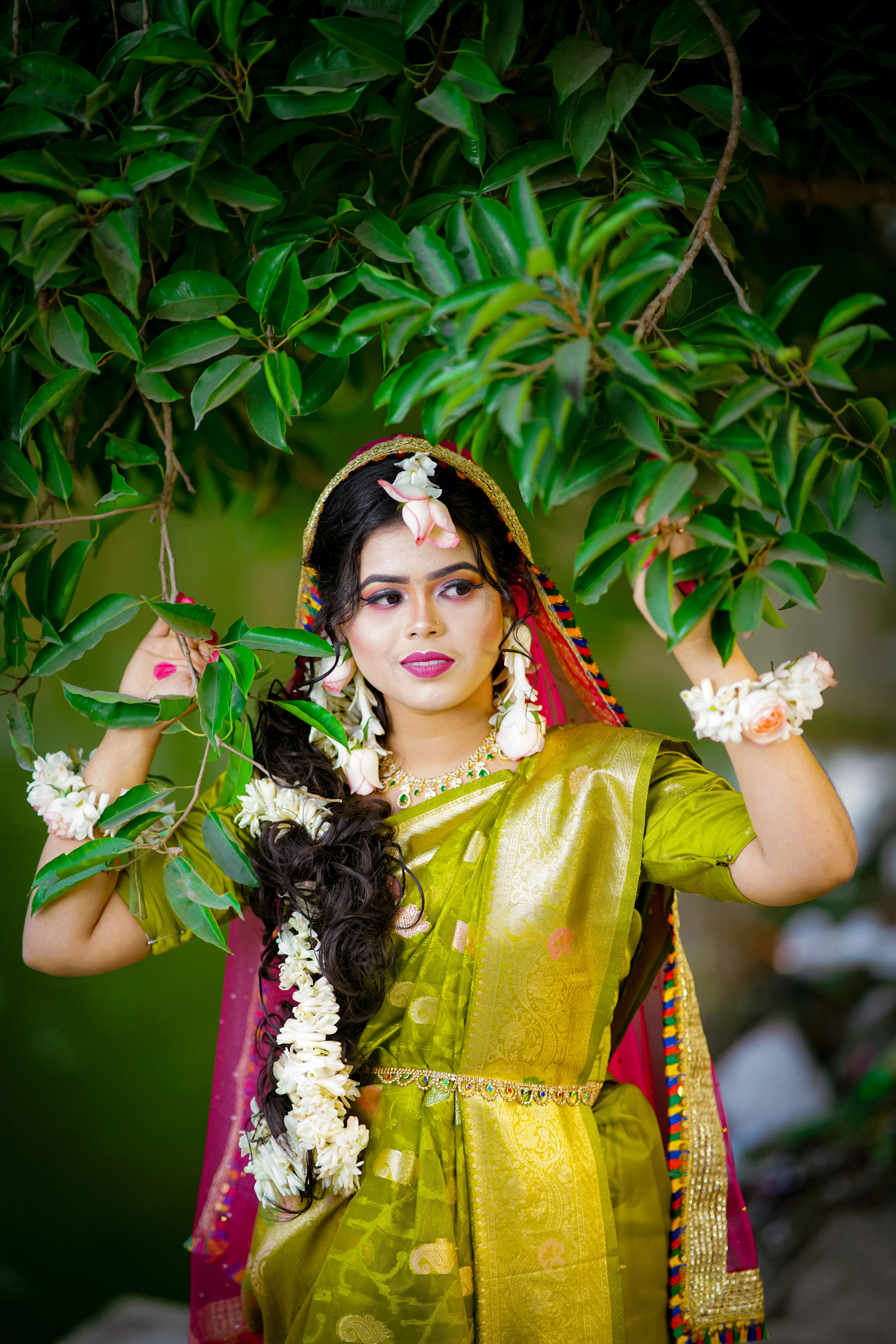 A woman poses in vibrant traditional attire adorned with floral accessories against lush foliage.