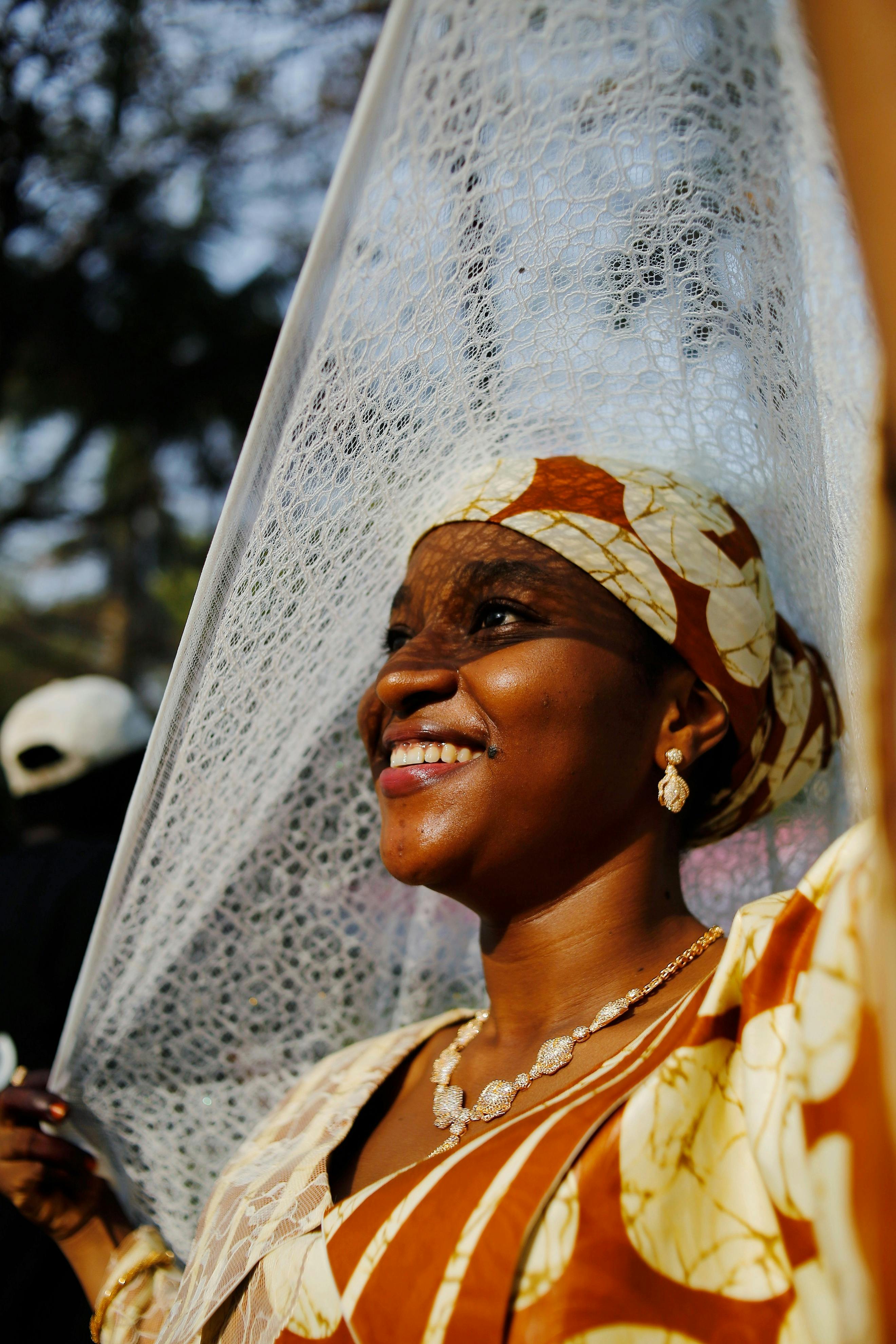 A woman in traditional Nigerian attire smiles under the sun in an outdoor setting in Abuja, Nigeria.