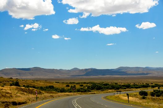 Open highway winding through a scenic mountainous landscape under a clear blue sky and fluffy clouds.