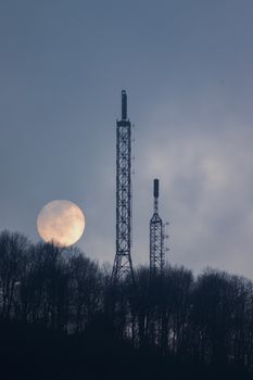 Moon rises behind communication towers above a silhouetted forest, creating a moody sky scene.