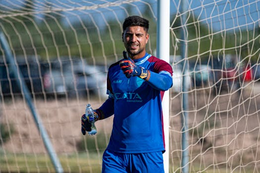 A goalkeeper in a blue jersey stands smiling by the goalpost outdoors.