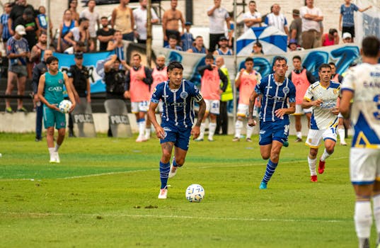 Players in blue and white jerseys compete in a lively soccer match with audience in the background.