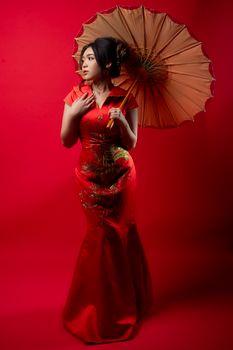 Asian woman in a cheongsam poses with a parasol against a red background indoors.