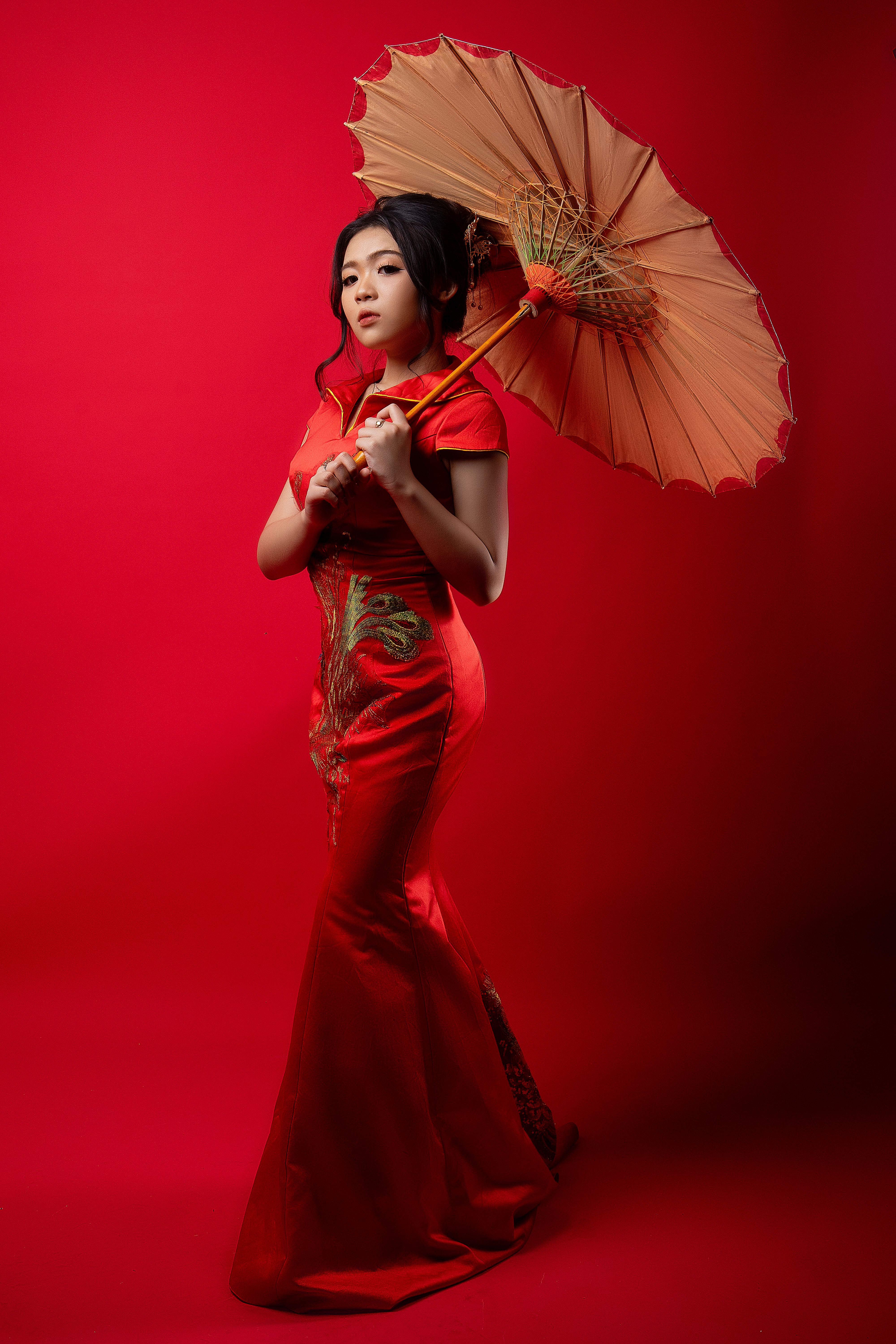 An elegant Asian woman poses in a red cheongsam holding a parasol against a red background.