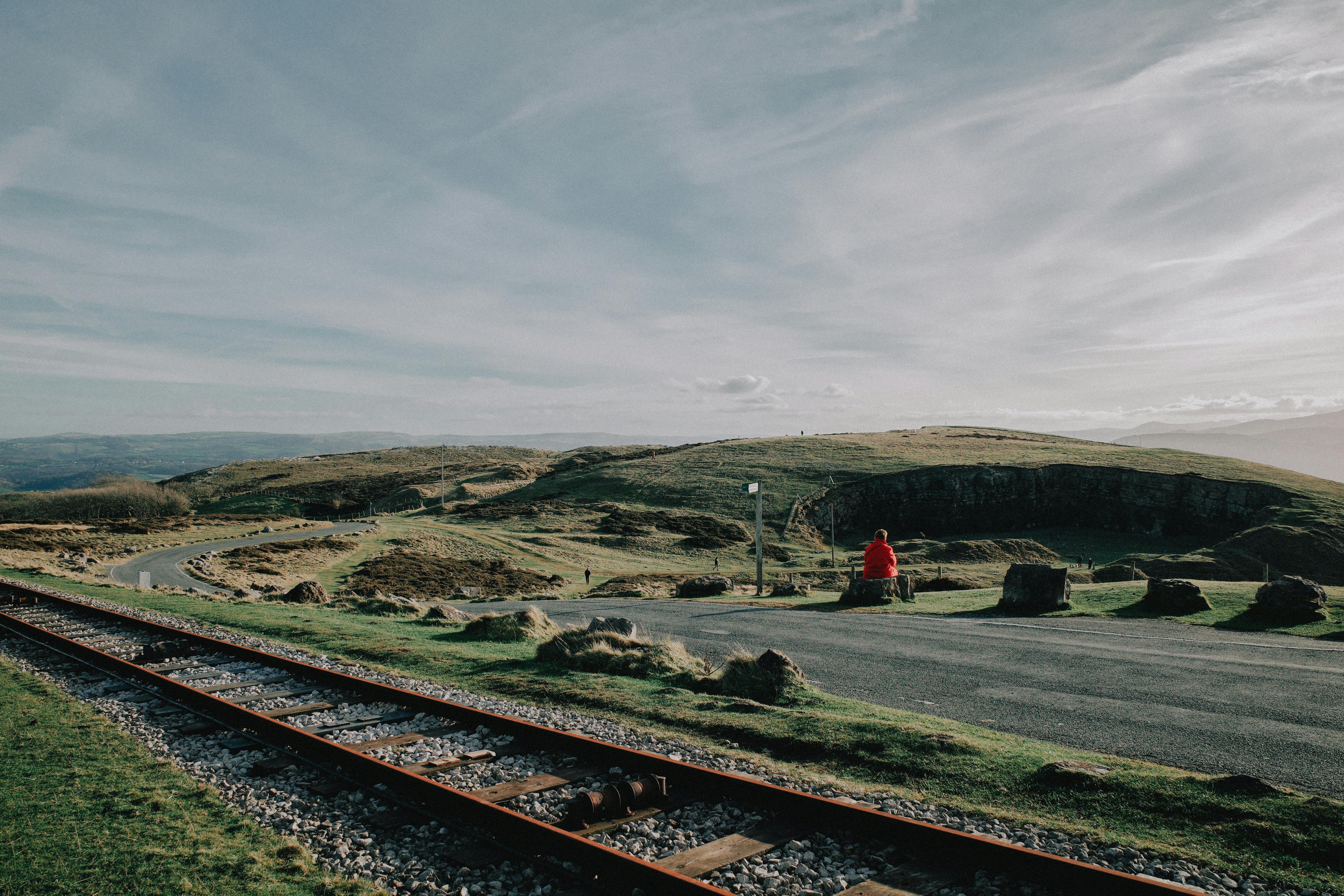 A lone figure in a red jacket observes a scenic railway landscape under a vast sky.