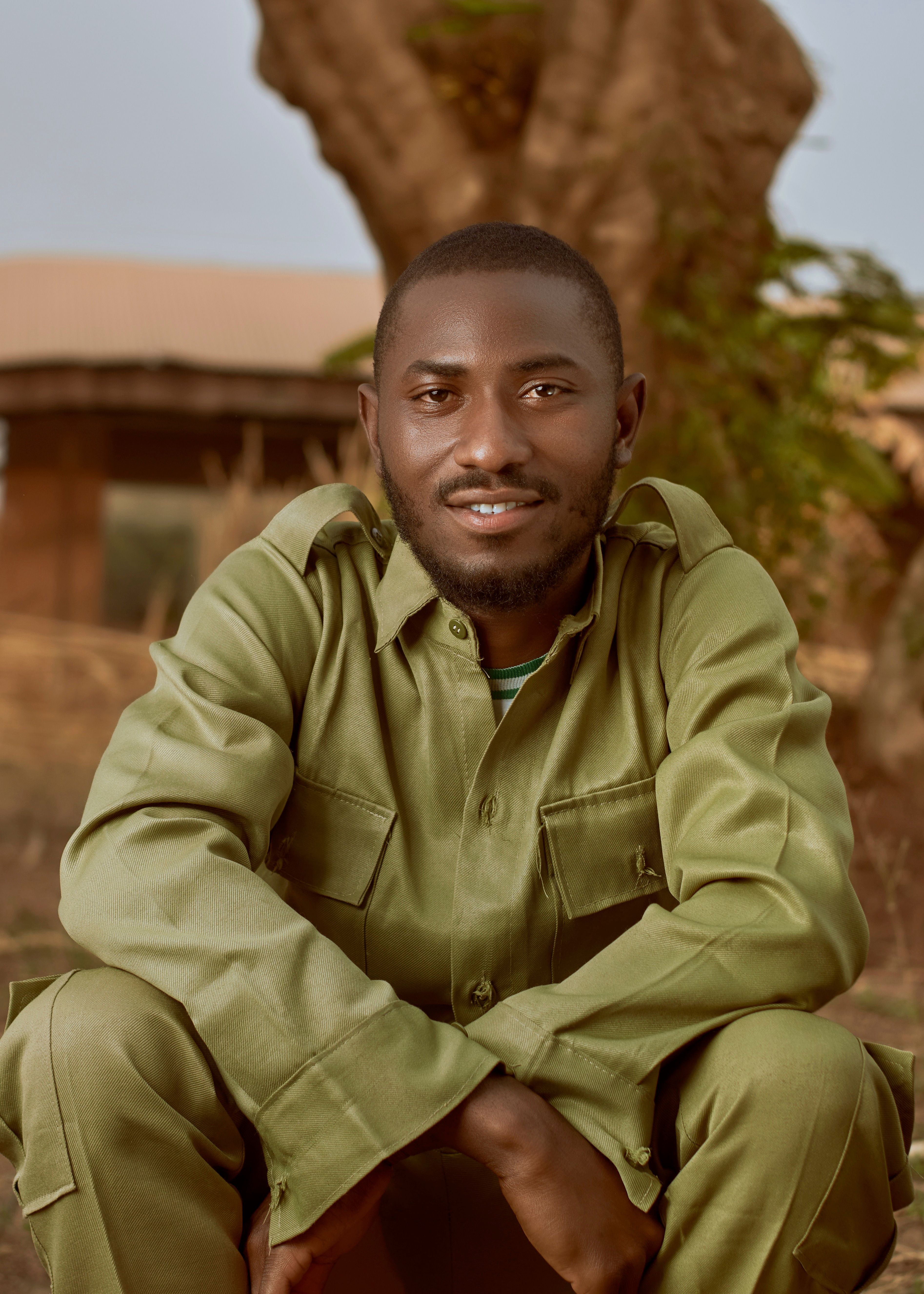 A young man in a green uniform smiles warmly, sitting outdoors with trees in the background.