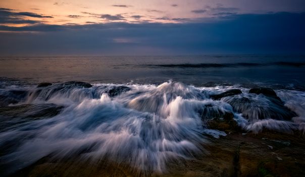 Waves crashing against rocks at sunset, capturing ocean's dramatic motion.