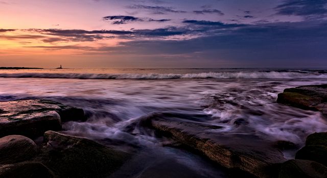 Stunning sunset over the ocean with waves crashing on rocky shoreline, creating a serene atmosphere.