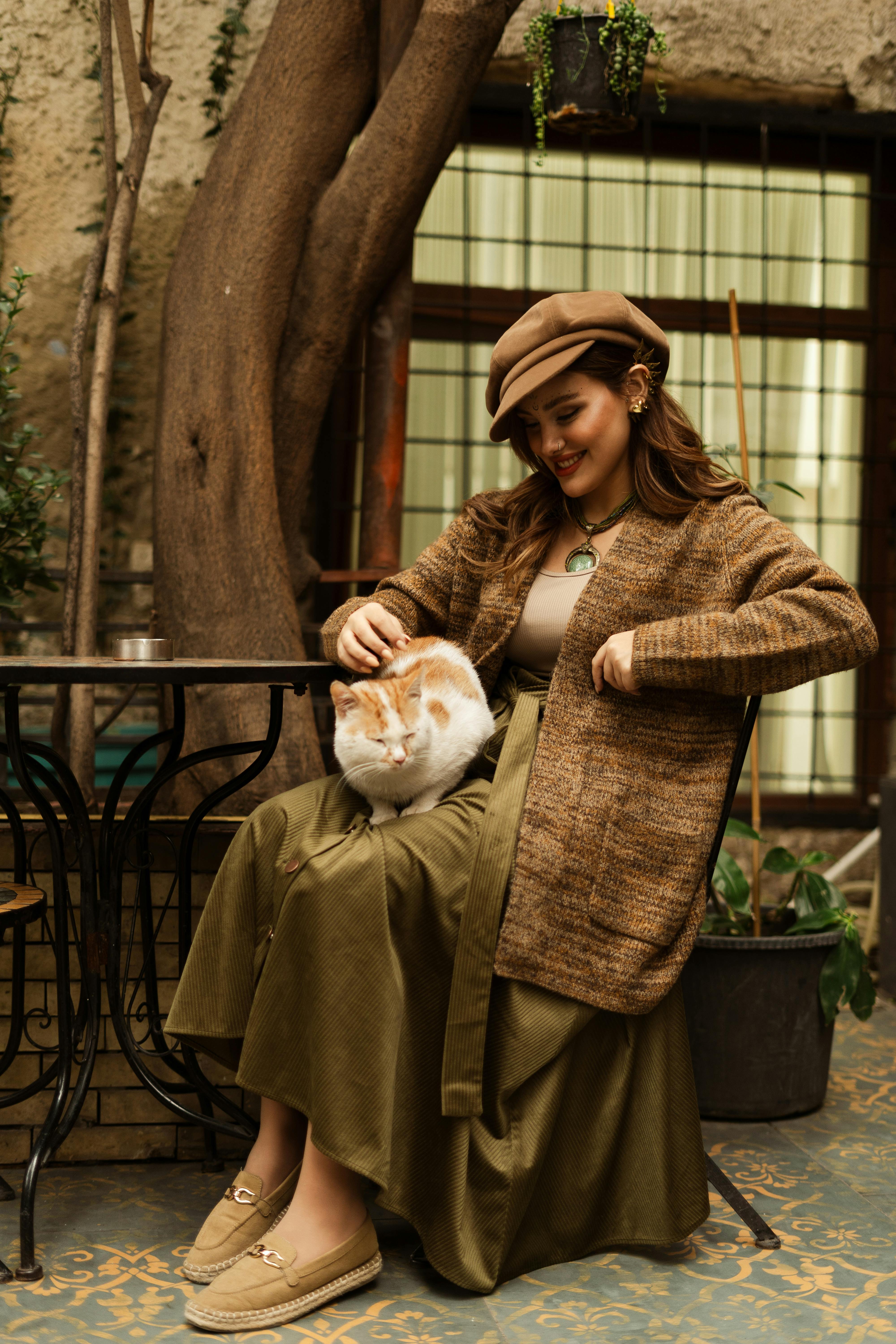 A fashionable woman in vintage attire sits with a cat in a rustic cafe setting.