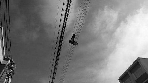 Monochrome image capturing shoes dangling from power lines in an urban setting.