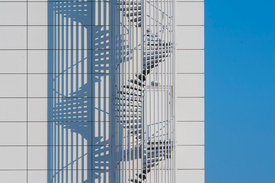 A sleek spiral staircase casts a geometric shadow on a building facade under a clear blue sky.