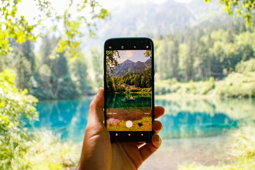 A hand holds a smartphone capturing a scenic mountain lake view with lush greenery.