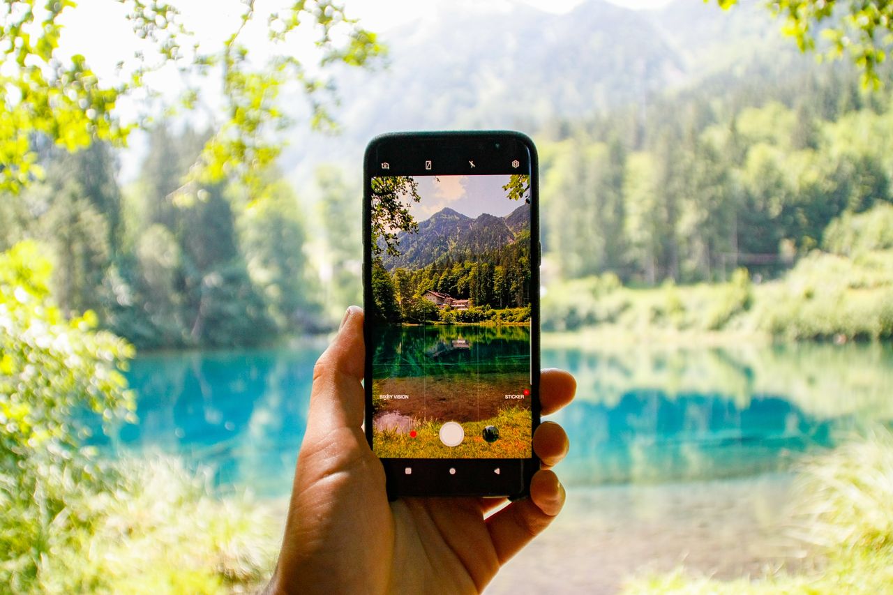 a hand holds a smartphone capturing a scenic mountain lake view with l