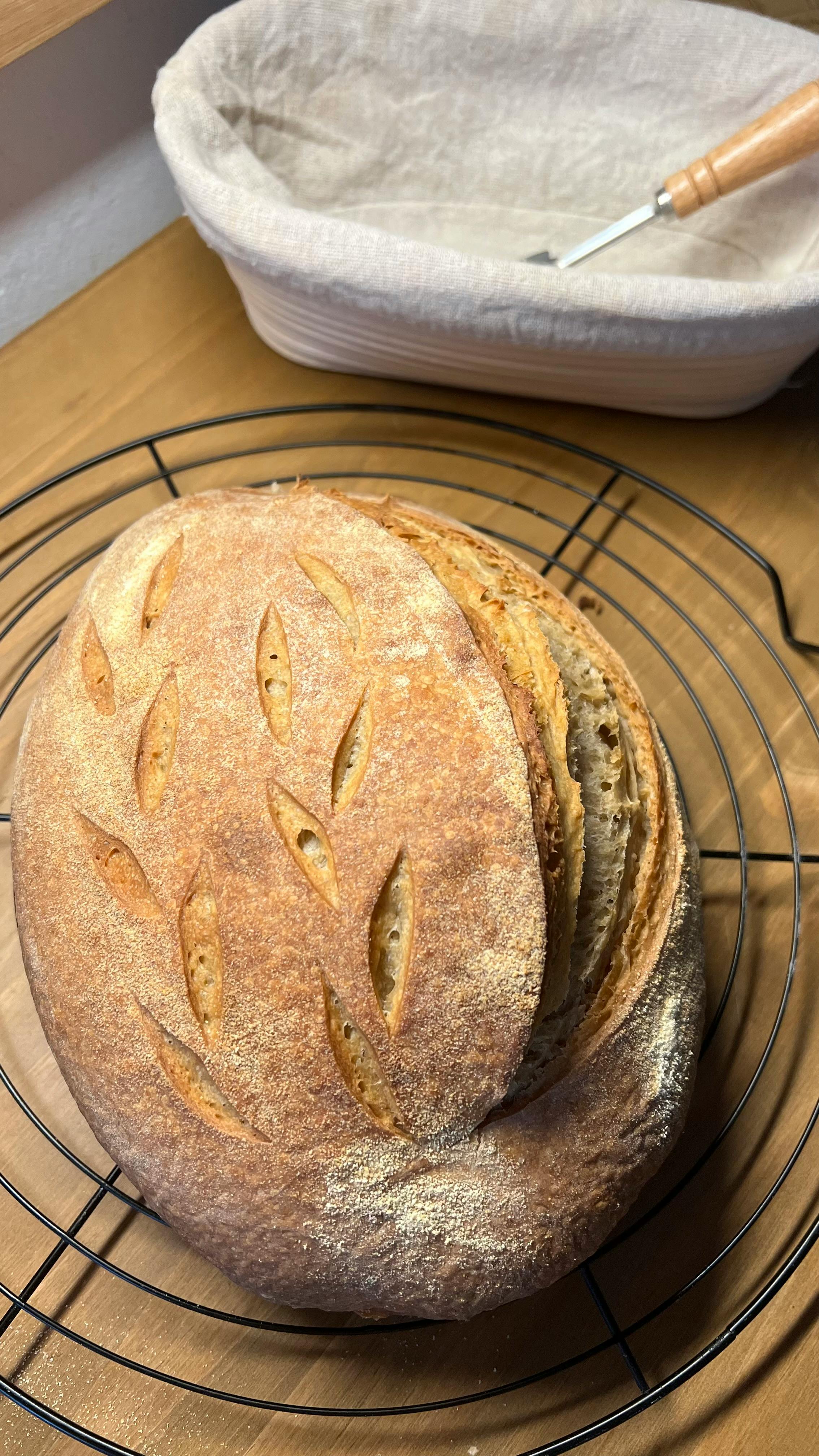 Golden brown sourdough bread with leaf patterns cooling beside a proofing basket.