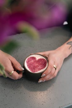 A latte with a heart design in frothed milk held by hands with rings on a concrete table.