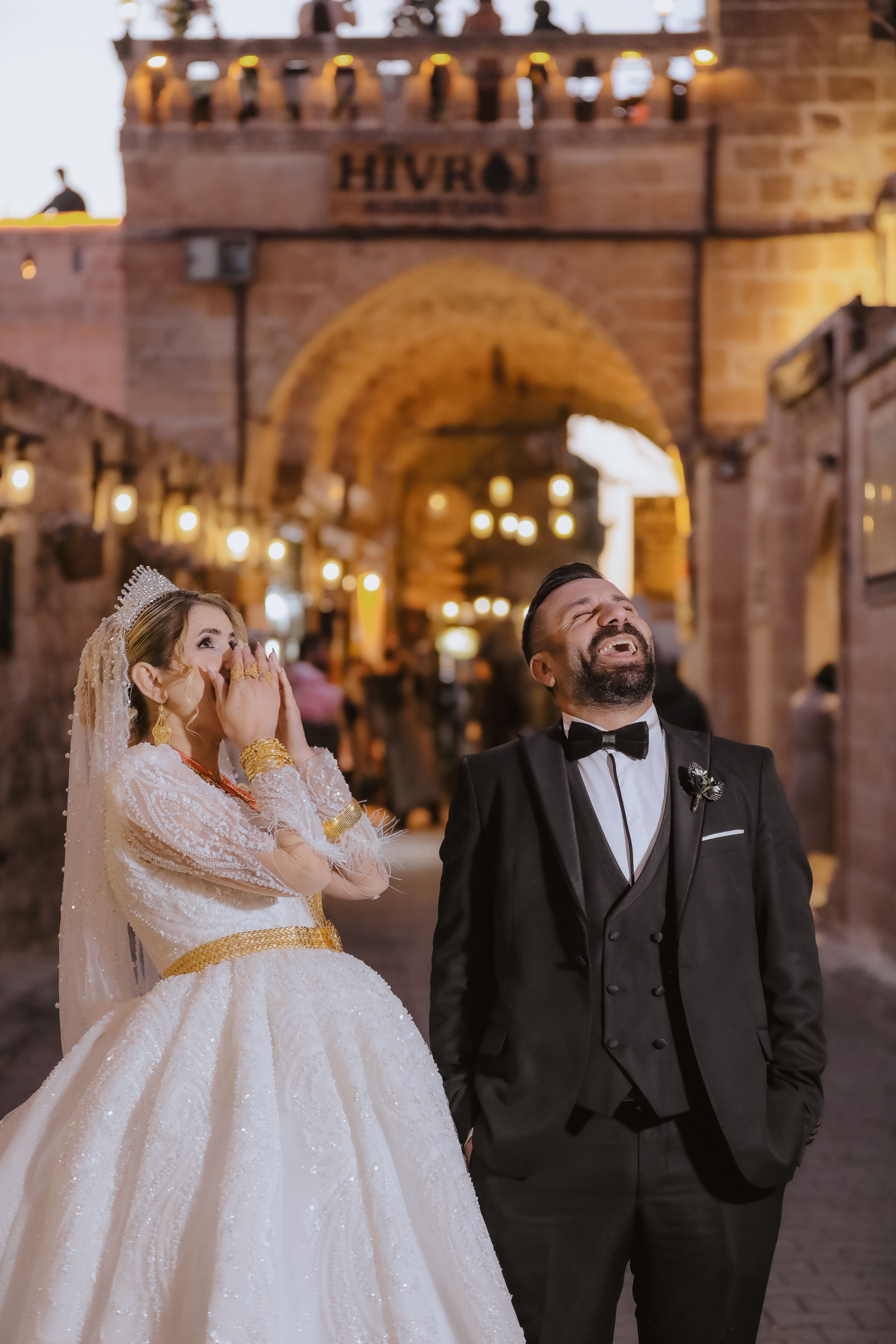 Laughing bride and groom under historic archway at night, celebrating love.