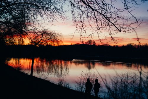 Free Two silhouettes by a serene lake during a vibrant sunset with a dramatic sky. Stock Photo