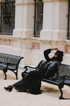 一位身穿黑色服装、头戴帽子的时尚女士在公园长凳上摆出姿势，展现现代时尚