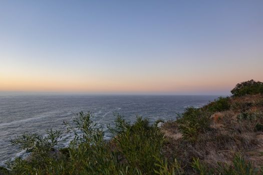 Tranquil ocean scene at sunset with a view from a coastal cliff, featuring calm seas and clear skies.