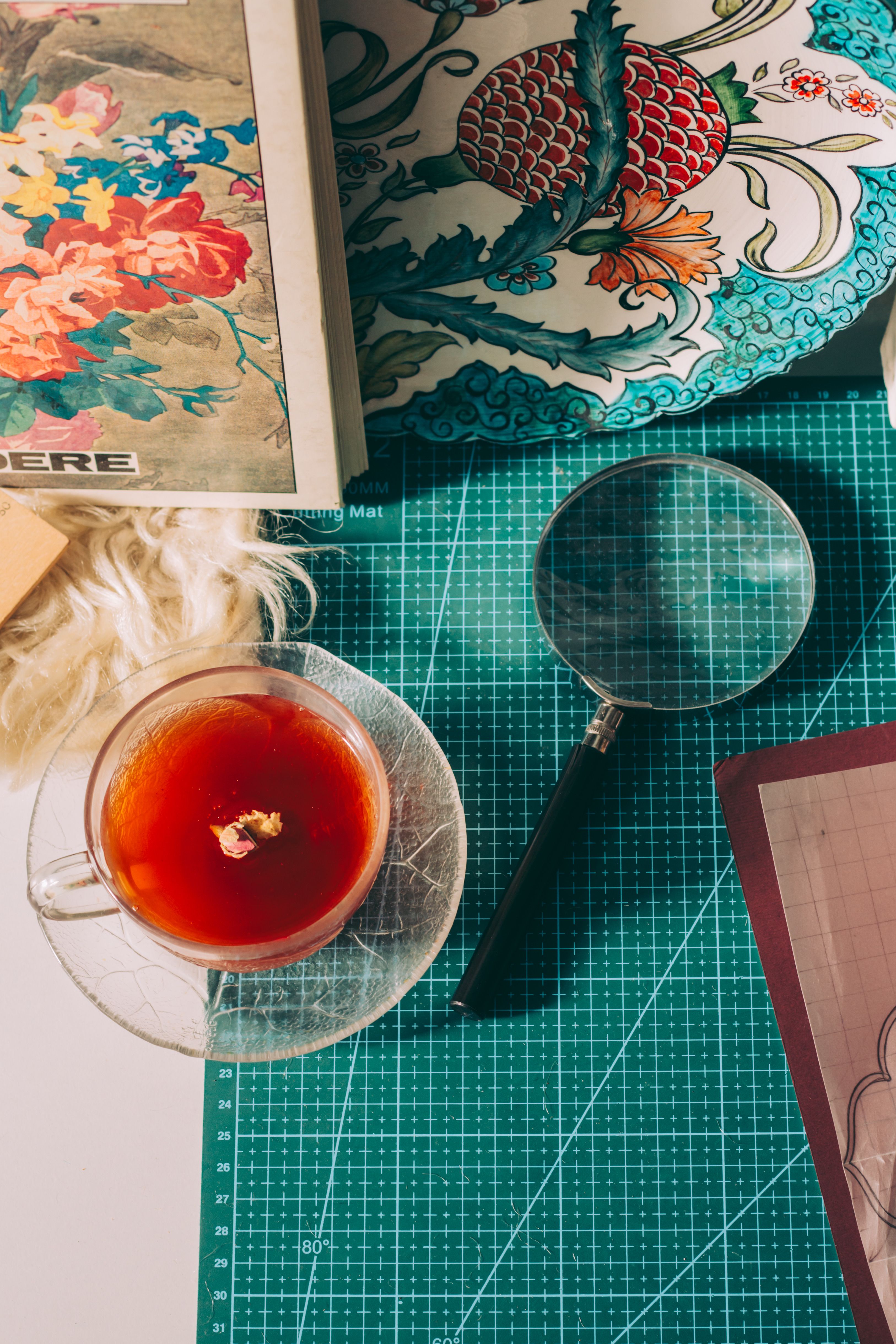 Flat lay of art materials, tea, and magnifying glass on cutting mat. Cozy and creative setup.