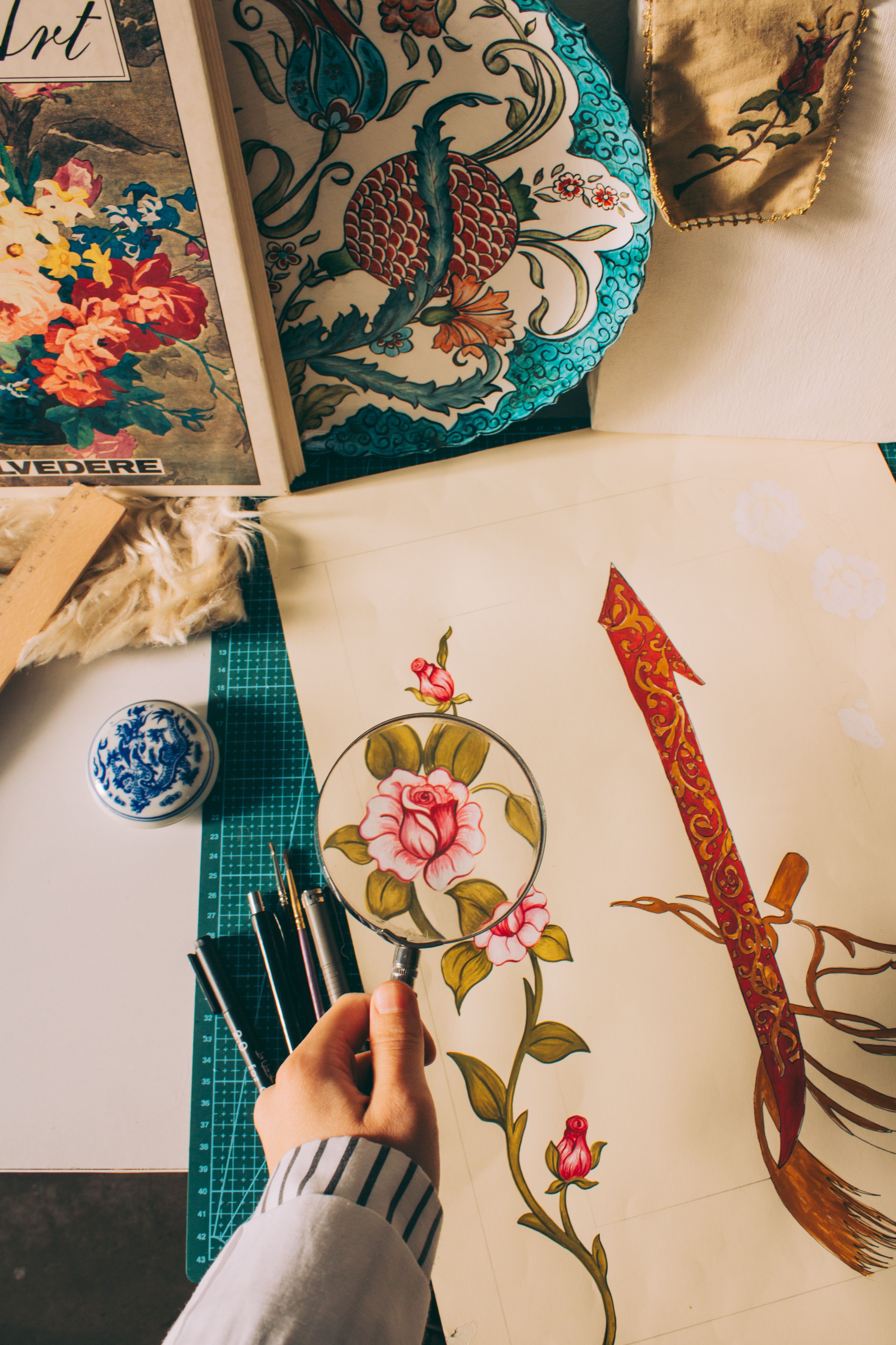 A creative workspace featuring a hand holding a magnifying glass over a floral sketch, surrounded by art supplies.