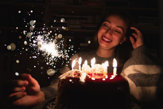 Smiling woman enjoying birthday cake with candles and sparkler indoors.