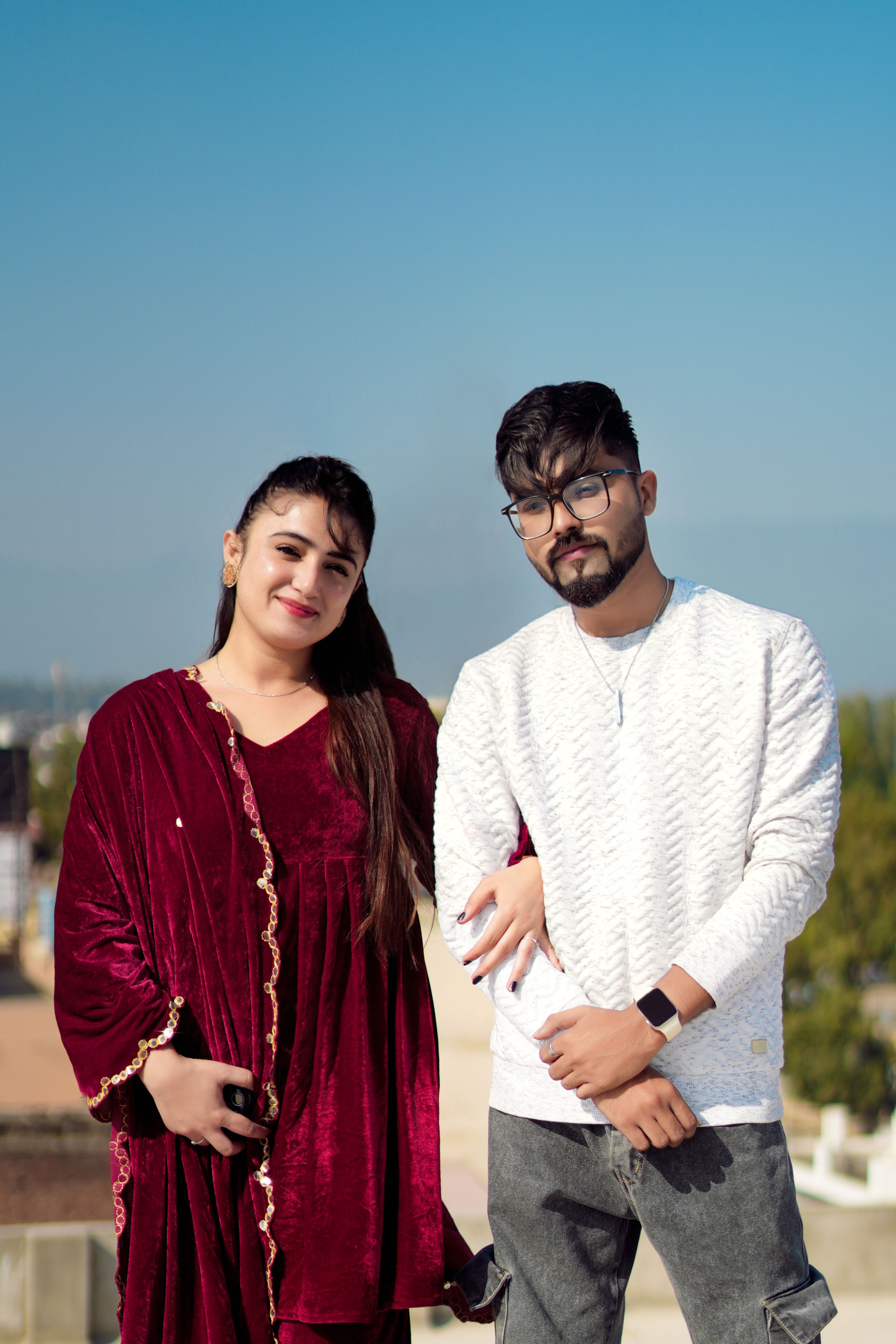 Young couple posing outdoors with clear blue sky background, showing modern fashion and style.