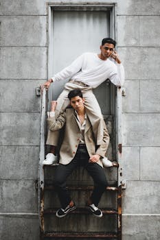 Two male models posing stylishly on a rusty staircase in an urban setting.