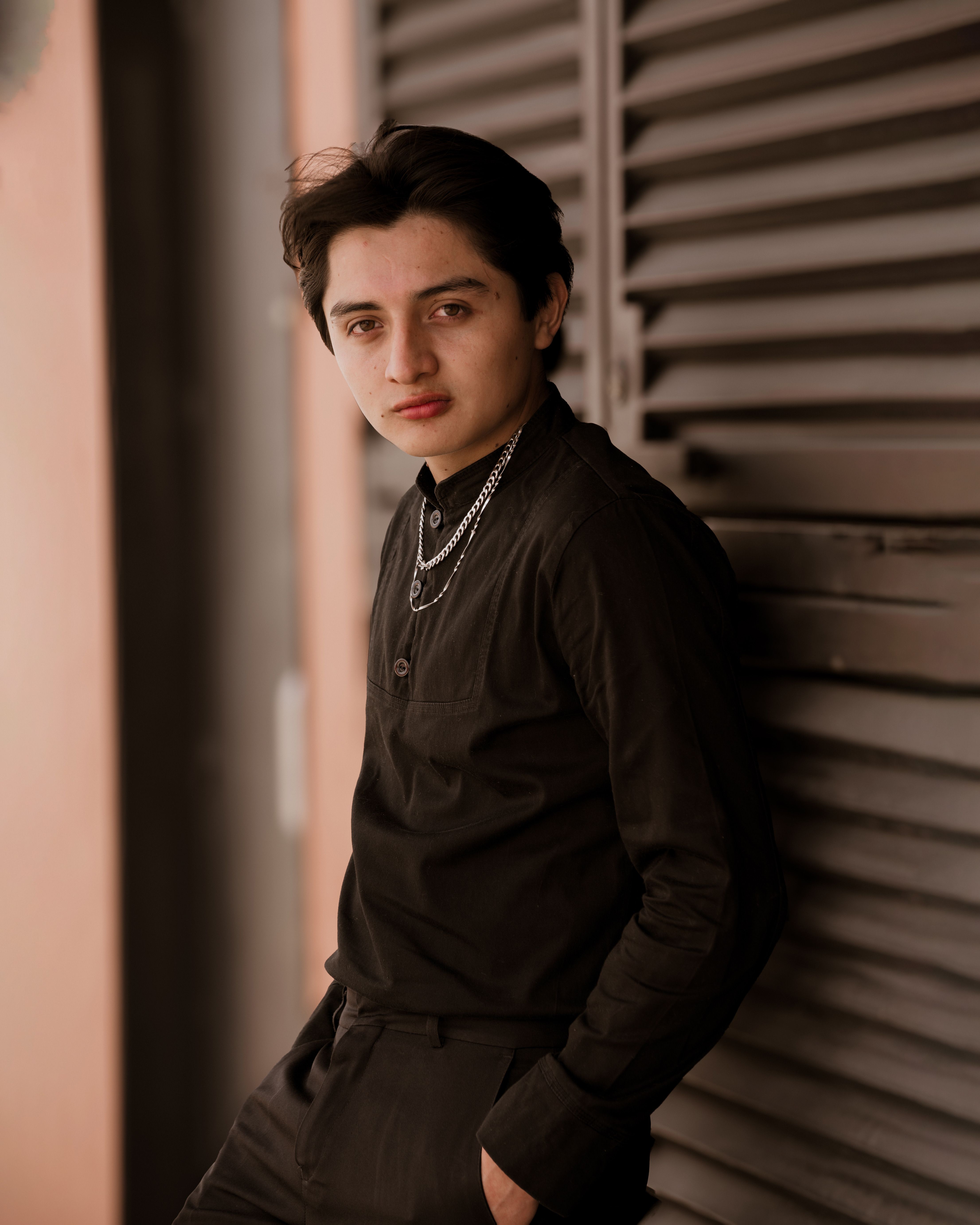Young adult in black attire leaning against shutters, wearing accessories, urban vibe.