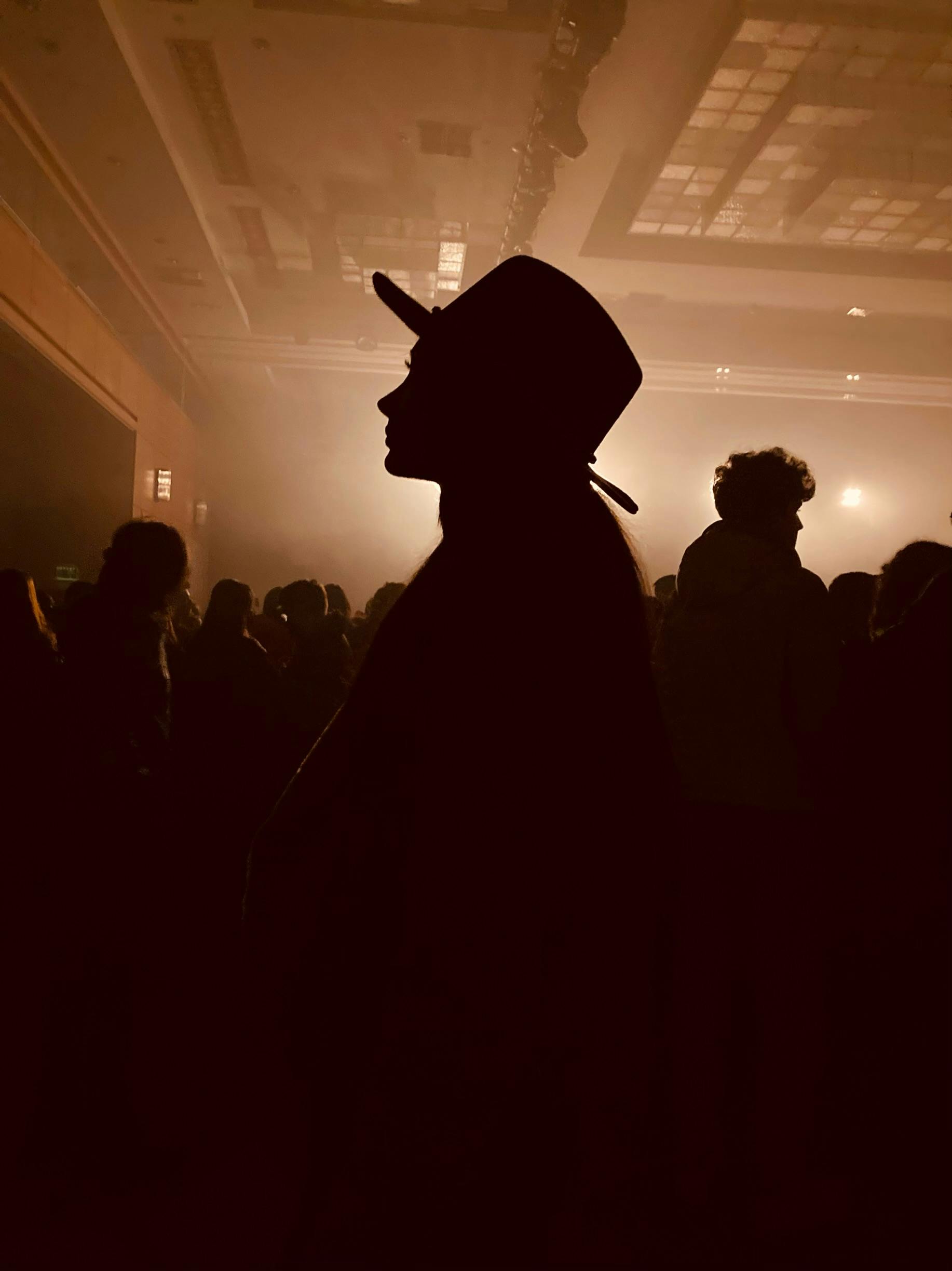Silhouette of a man wearing a hat at an indoor concert with atmospheric lighting.