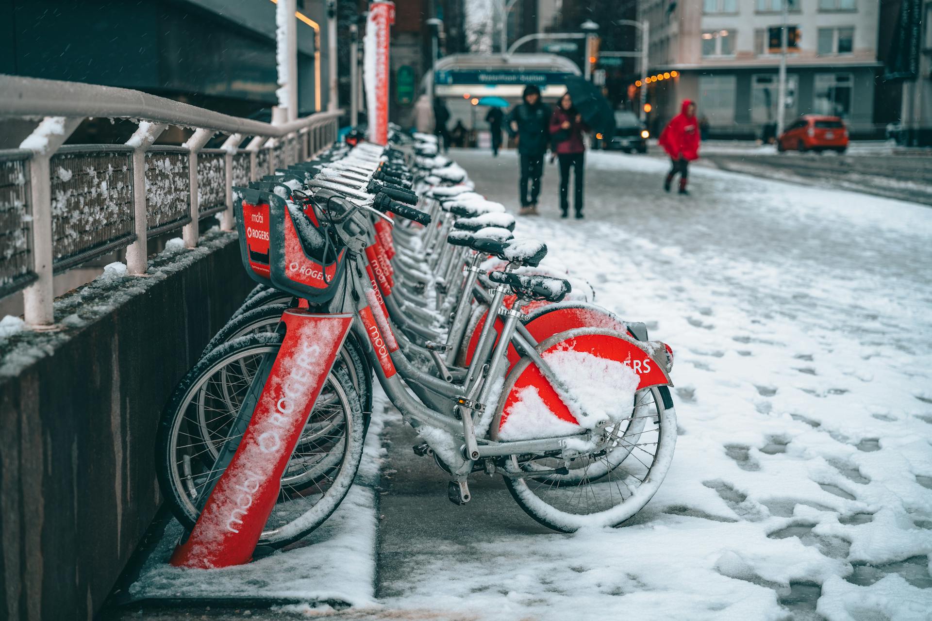 Bicycles covered in snow at a city rack during winter, people walking nearby.