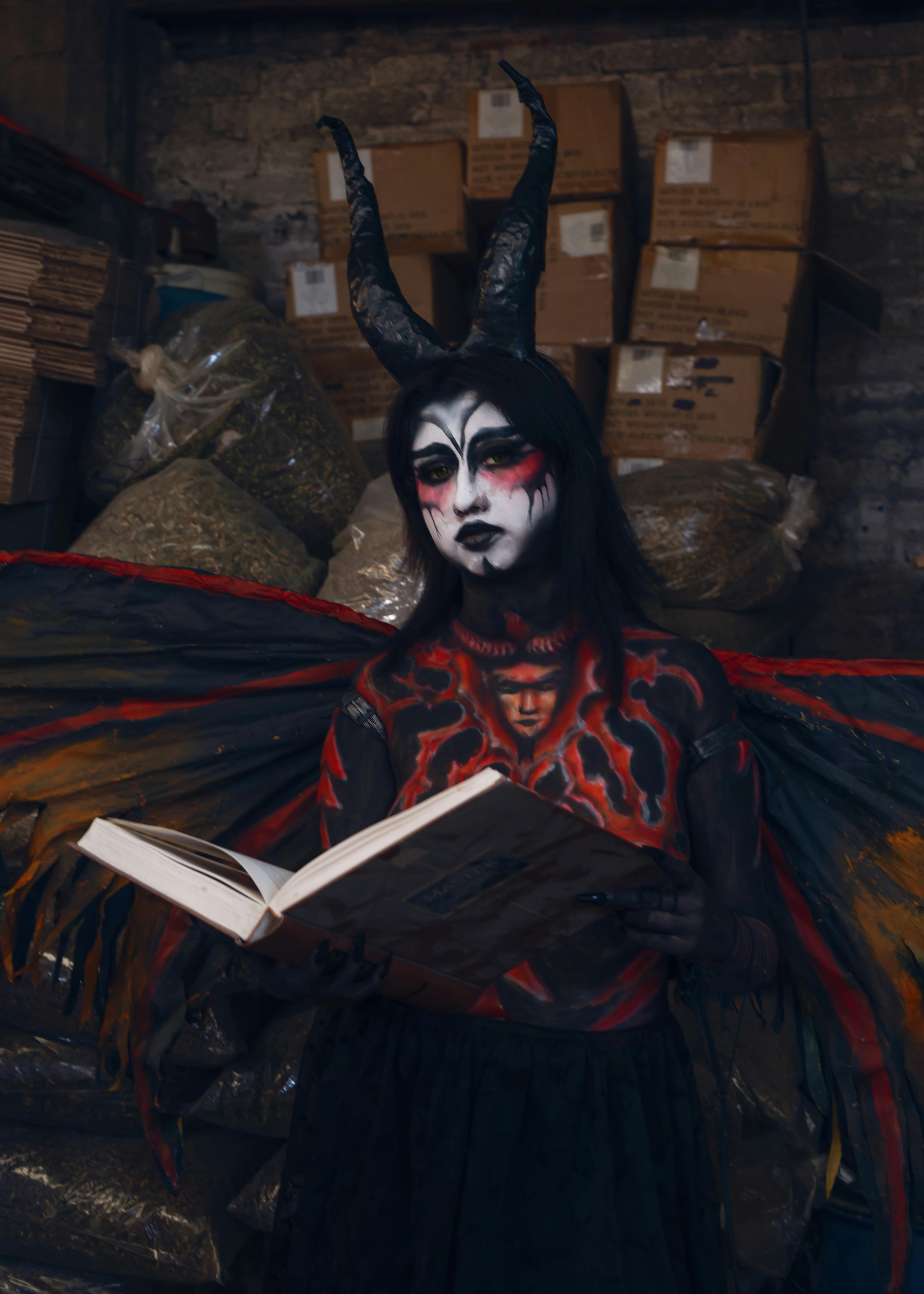 grátis Pessoa em traje demoníaco com chifres e pintura facial segurando um livro, em ambiente interno. Foto profissional