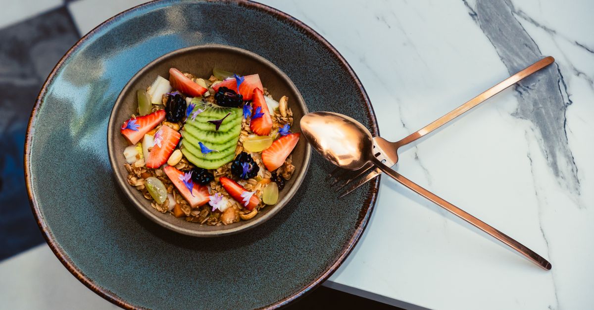 Tigela vibrante de frutas e granola servida com talheres elegantes sobre uma superfície de mármore.