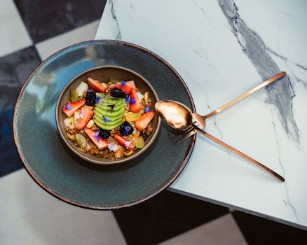 Vibrant fruit and granola bowl served with elegant cutlery on a marble surface.