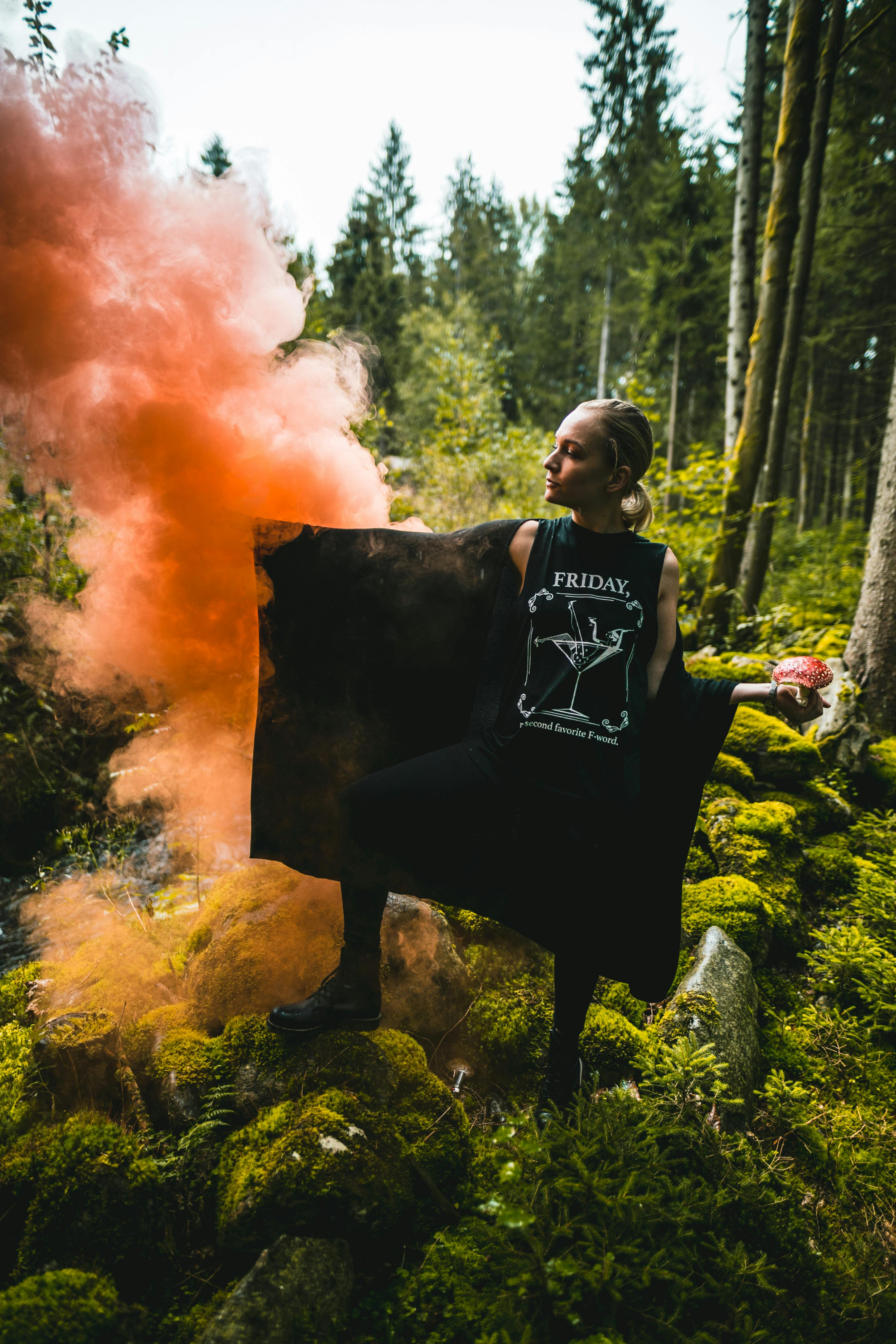 Woman in forest with red smoke bomb and mossy ground, creating a dramatic scene.