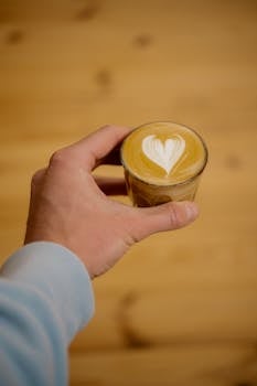 Close-up of a hand holding a cortado coffee with heart-shaped latte art on a wooden table.