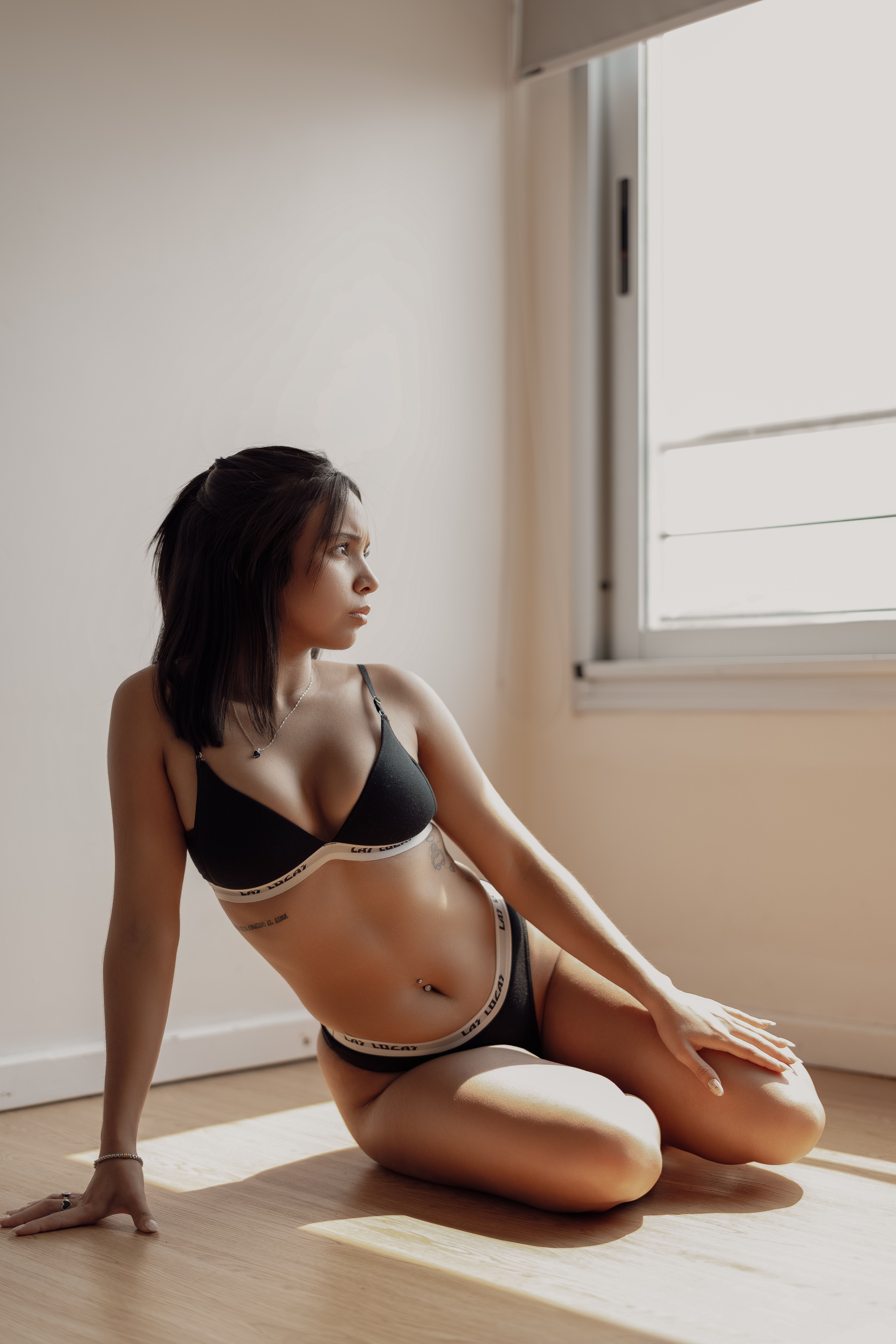 Young woman poses in black lingerie by a sunlit window in a minimalistic room.