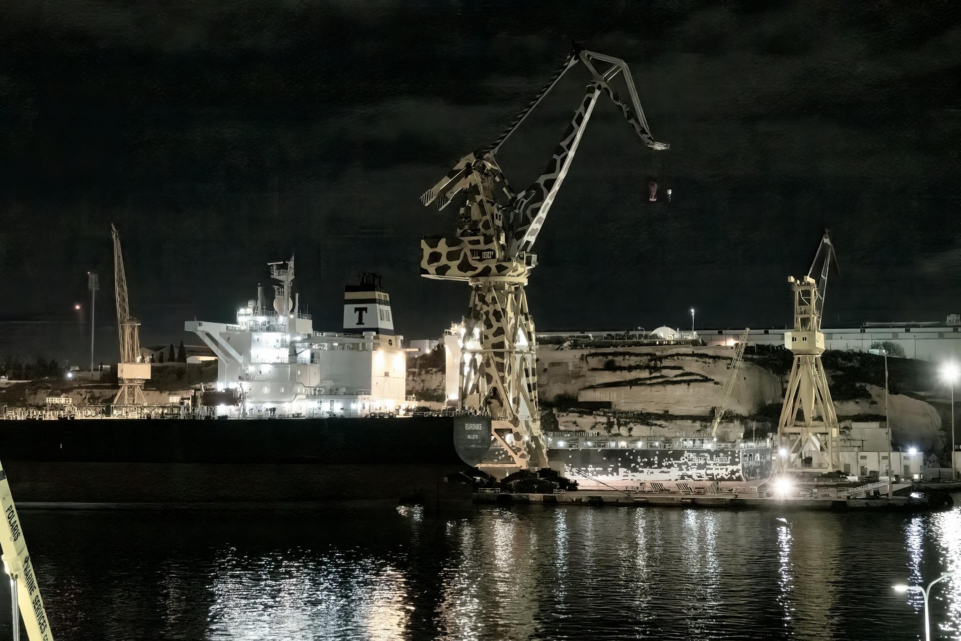 A ship docked at night at a shipyard in Malta with illuminated cranes casting reflections.