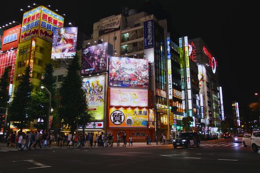 Bustling street scene in Akihabara, Tokyo at night with bright neon signs and animated billboards.