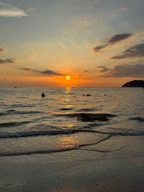 Free Serene beach sunset with people swimming in tranquil waters and a vibrant sky. Stock Photo