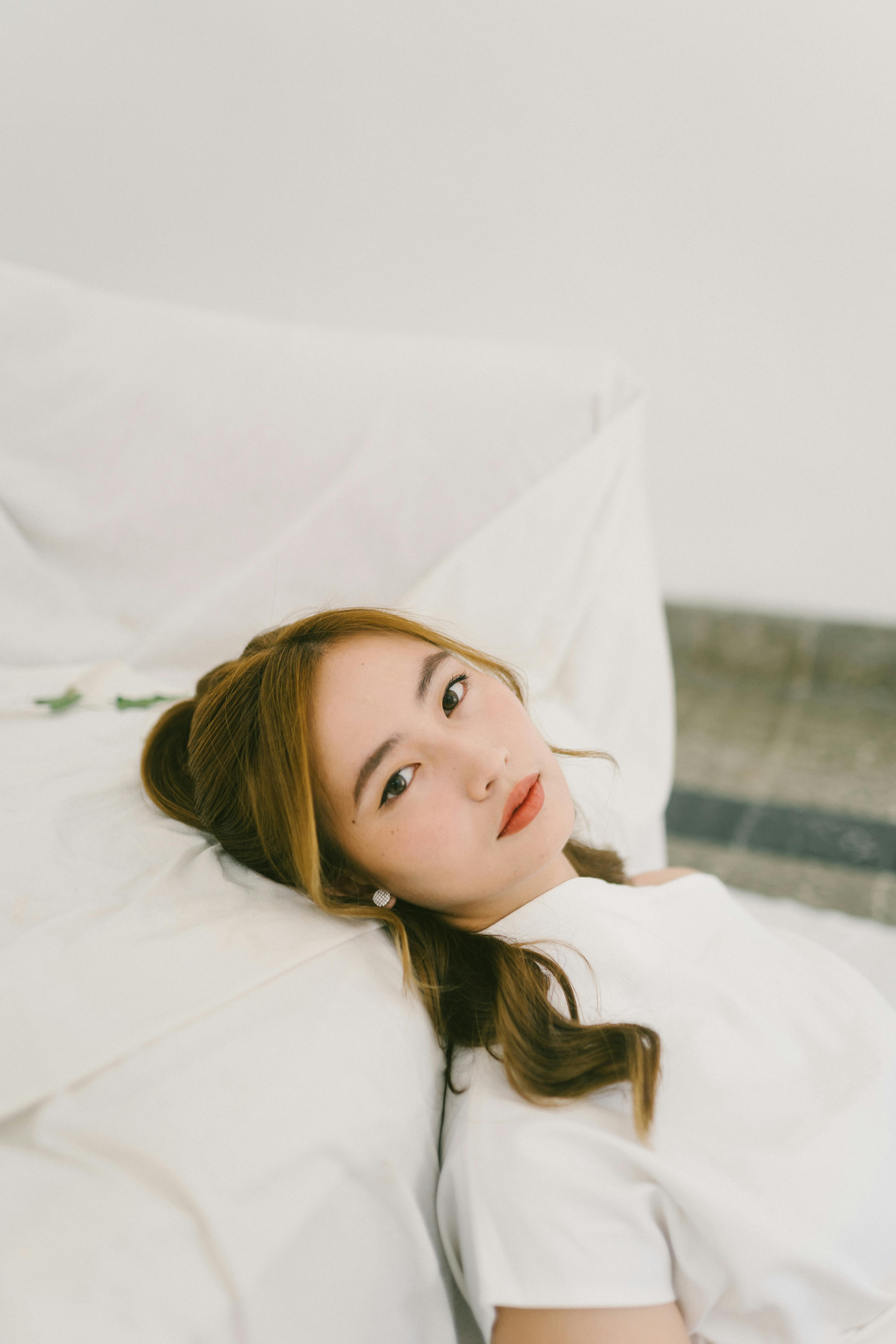 Portrait of a woman lying on a white bed in a serene setting. Soft lighting enhances the relaxed mood.