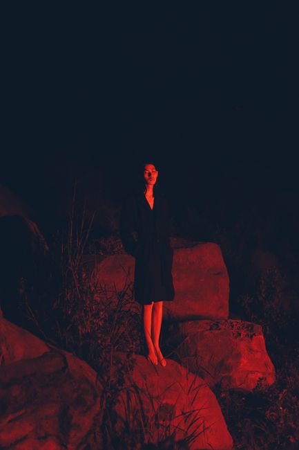 A mysterious woman stands on rocks, illuminated by red light at night.
