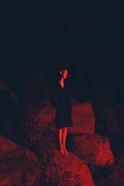 A contemplative woman stands illuminated by red light on rocks at night.