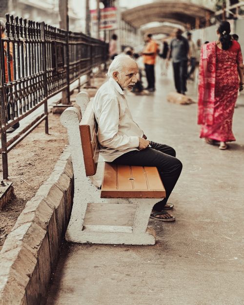 Free Elderly man in white shirt sitting on a street bench, pondering in a bustling outdoor setting. Stock Photo