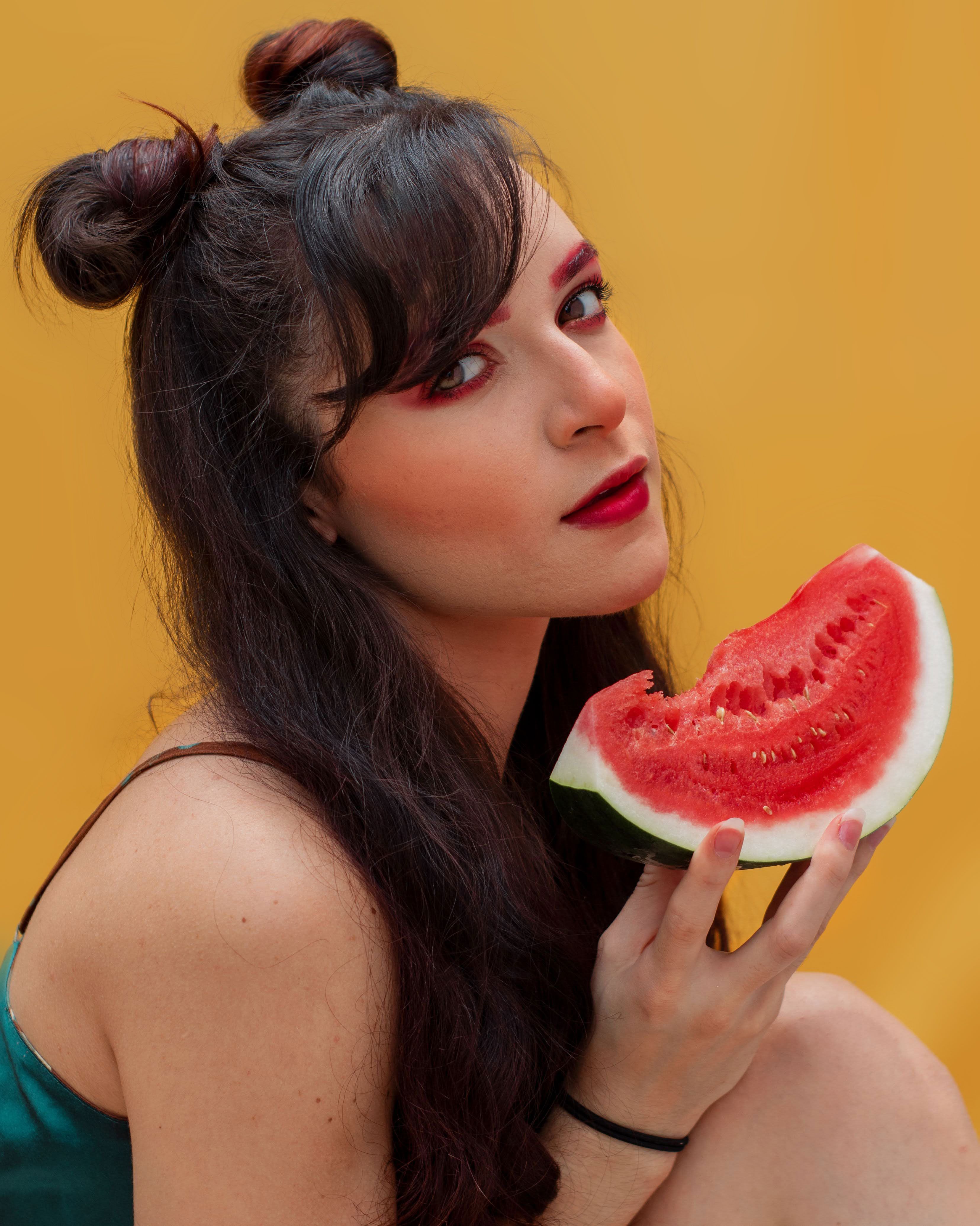 Stylish young woman with bold makeup holding a slice of watermelon against a bright backdrop.