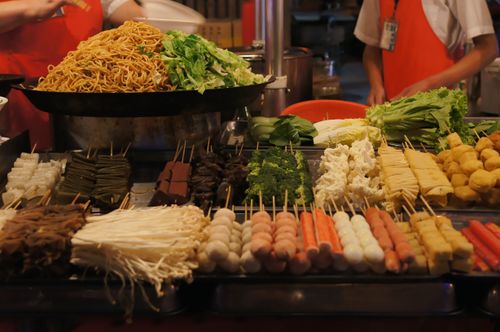 Free Colorful Asian street food display with assorted skewers and fresh vegetables under warm lighting. Stock Photo
